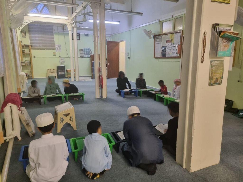 Children reading Qur'an on rehals in small circles across the prayer hall floor